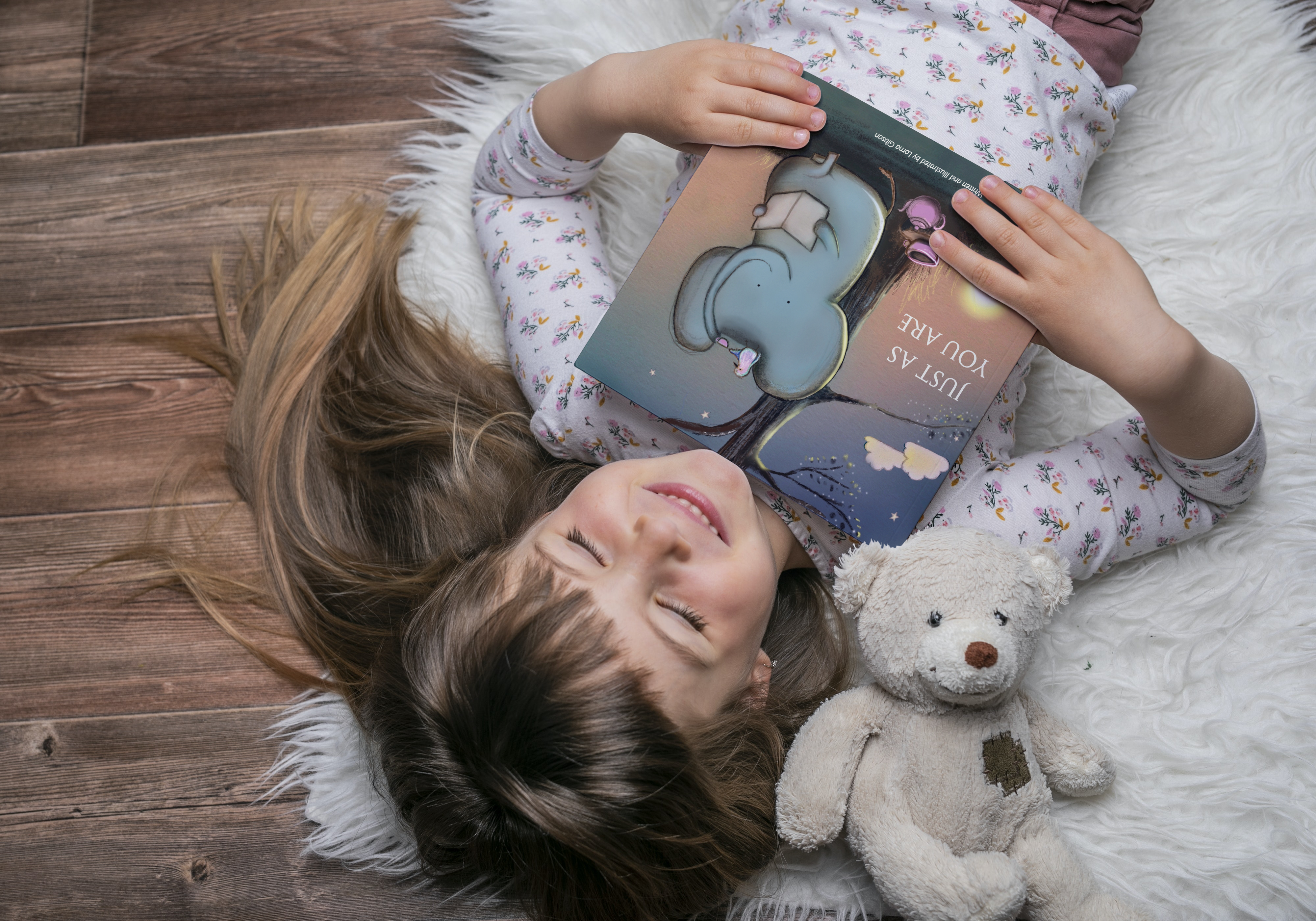 Gift ideas for kids - Image shows a little girl holding a copy of Just As You Are beside her teddy bear lying on a cosy fluffy rug